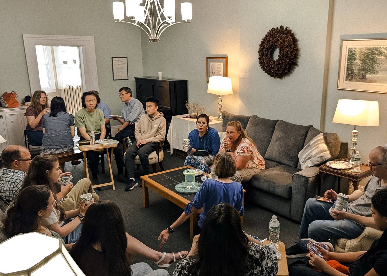 A living room full of people of different ages and countries, sitting in a circle, eating and talking.