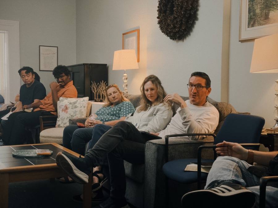 People sitting on sofas and armchairs in a home, listening to someone talk.