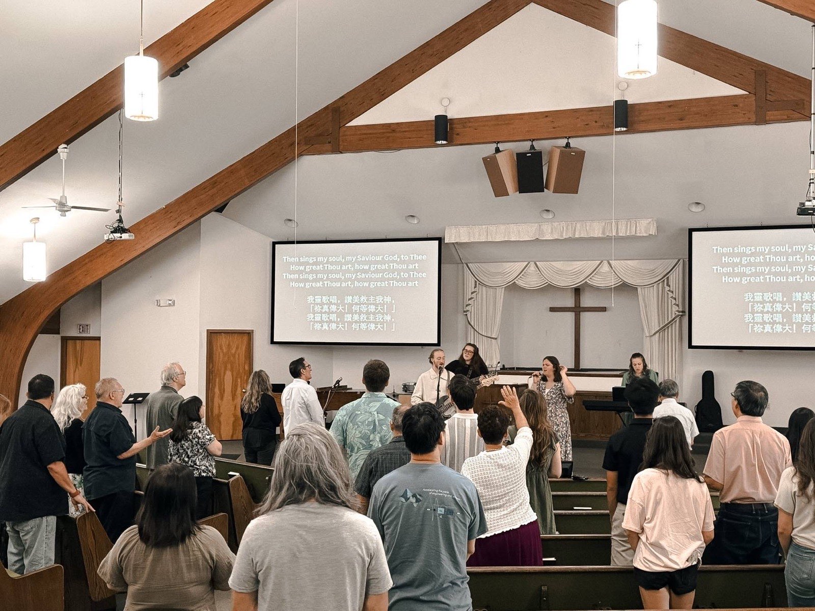 A room of people standing to sing, with the song words on the screen in both English and Chinese.
